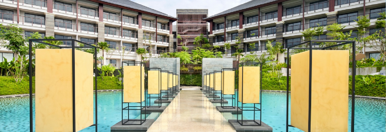 Marriott resort exterior in Bali featuring a stone walkway crossing a turquoise pool, flanked by rows of tall, yellow rectangular lanterns in black metal frames.