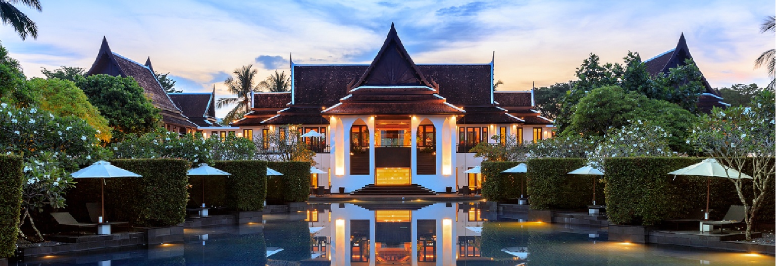 A luxury Marriott resort in Khao-Lak with traditional Thai-style architecture, featuring a large swimming pool in the foreground, surrounded by hedges and white umbrellas.