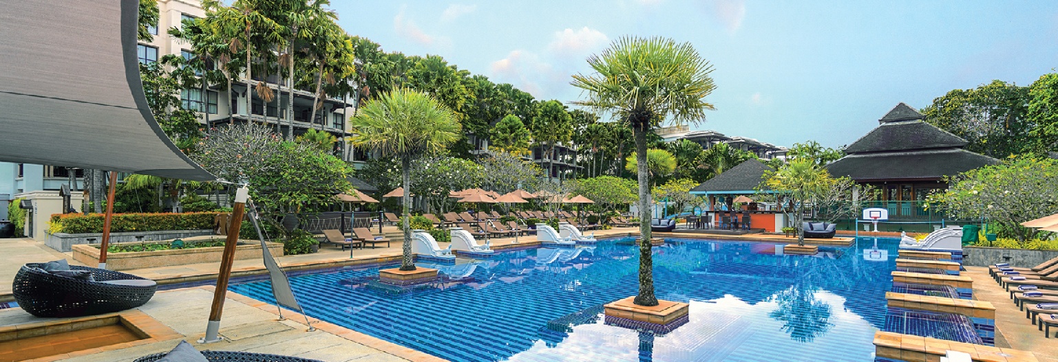 A large, rectangular swimming pool with blue tiles, surrounded by palm trees, lounge chairs, and resort buildings under a clear, bright blue sky at a Phuket Marriott resort.